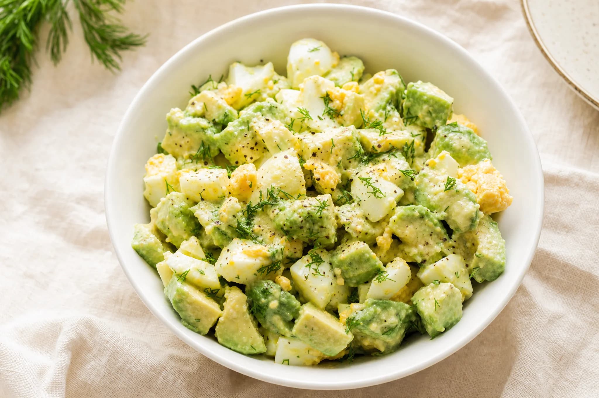 Creamy avocado egg salad in a white ceramic bowl with herbs on top