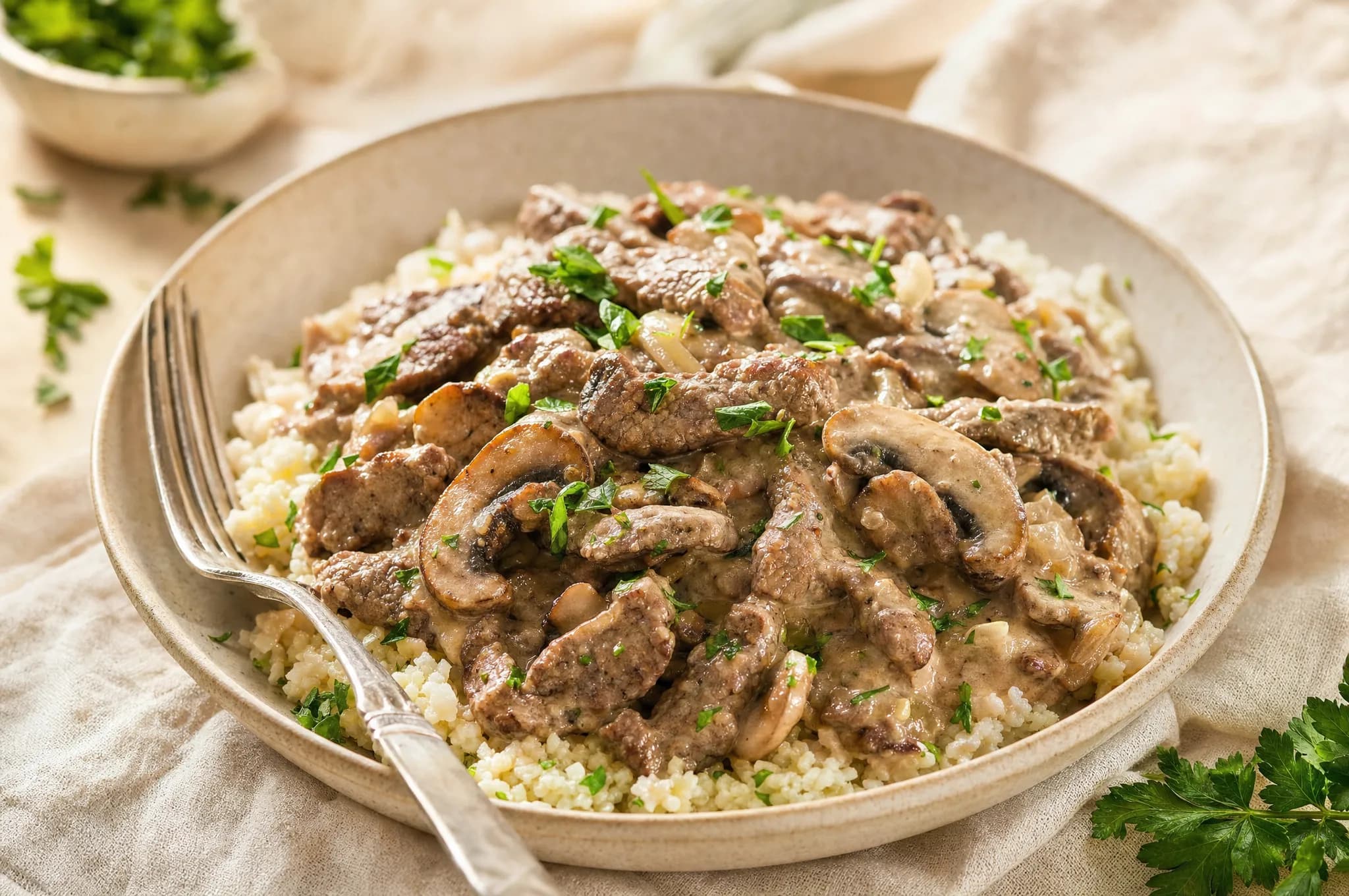 Strips of seared beef and sliced mushrooms in a creamy sauce over cauliflower rice in a shallow bowl, scattered with parsley, a fork resting alongside