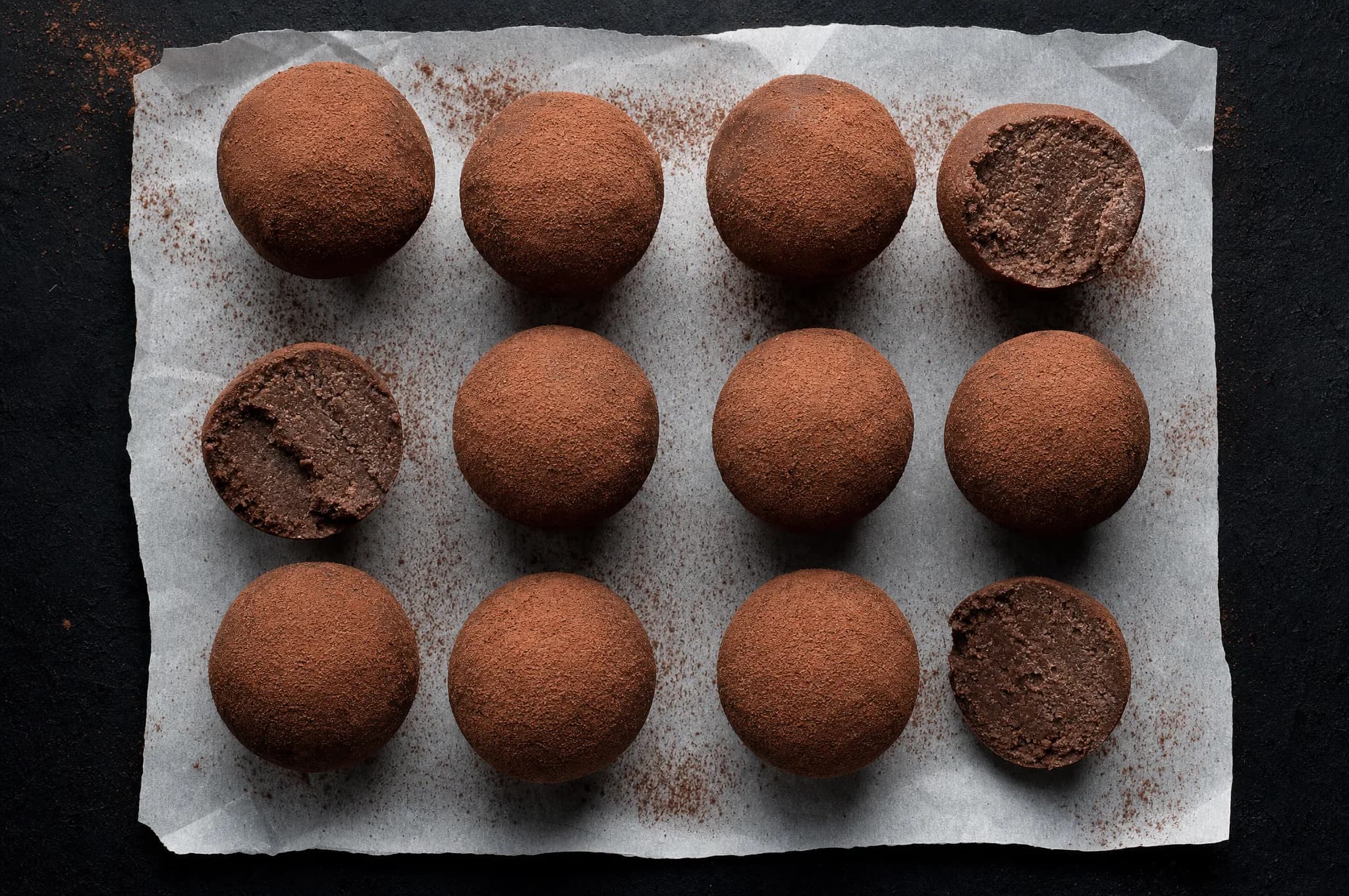 Twelve round keto chocolate peanut butter fat bombs dusted with cocoa powder, arranged on parchment paper with a dark background