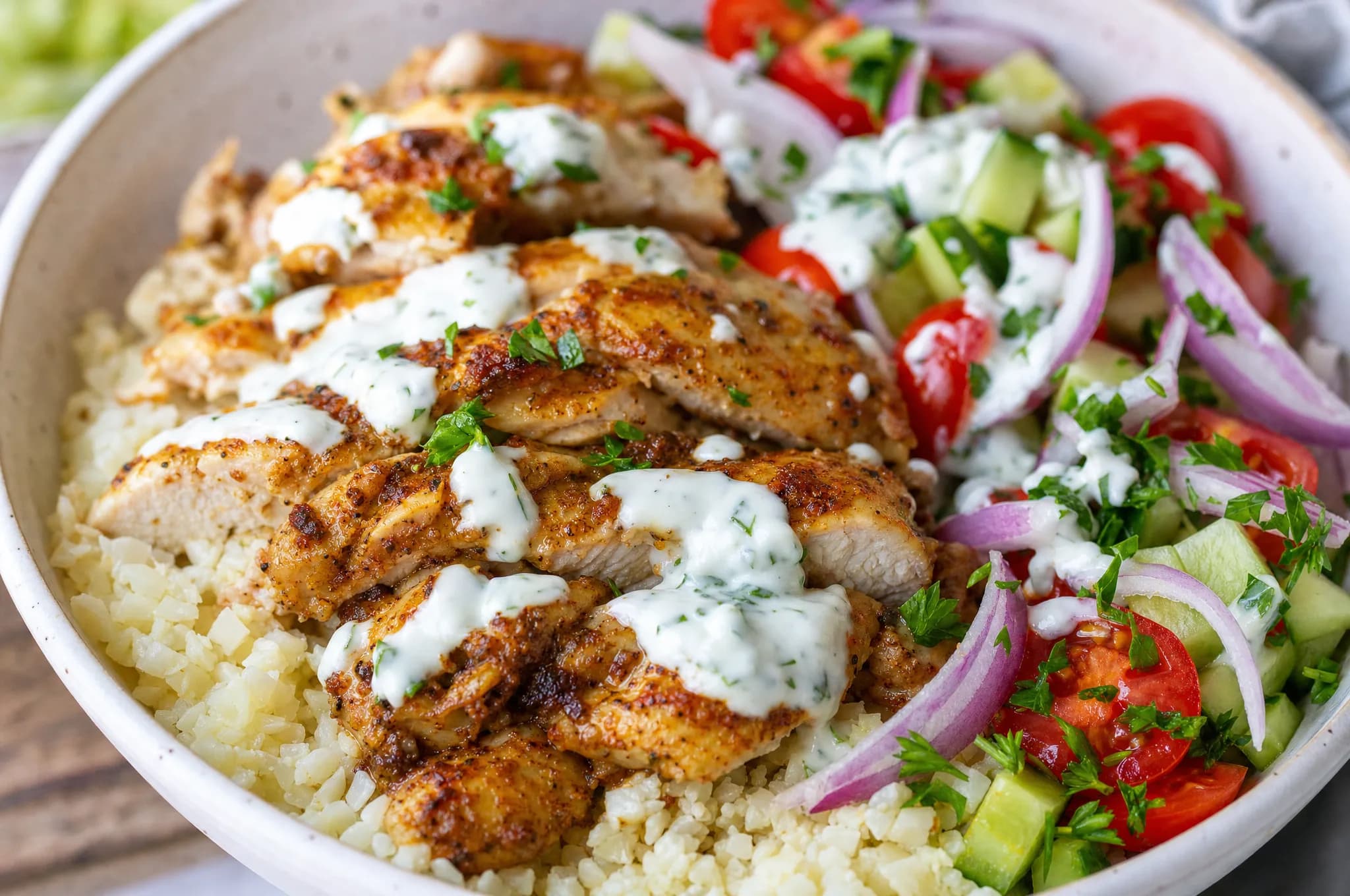 Four keto meal-prep bowls of sliced shawarma-spiced chicken thighs over cauliflower rice with garlic-yogurt sauce, cucumber, cherry tomato, red onion, and parsley