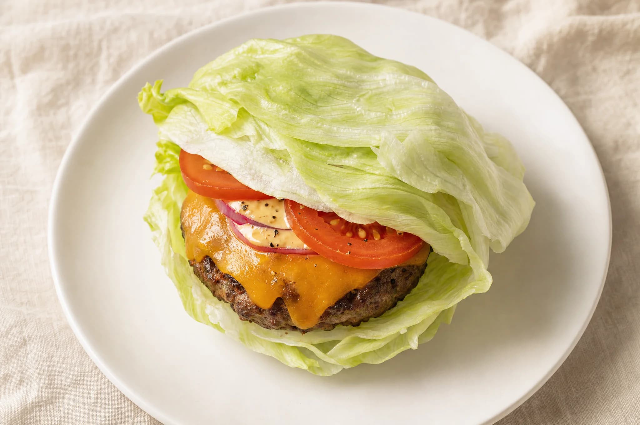 A keto burger in a crisp iceberg lettuce wrap on a white plate, beef patty with melted cheese and tomato