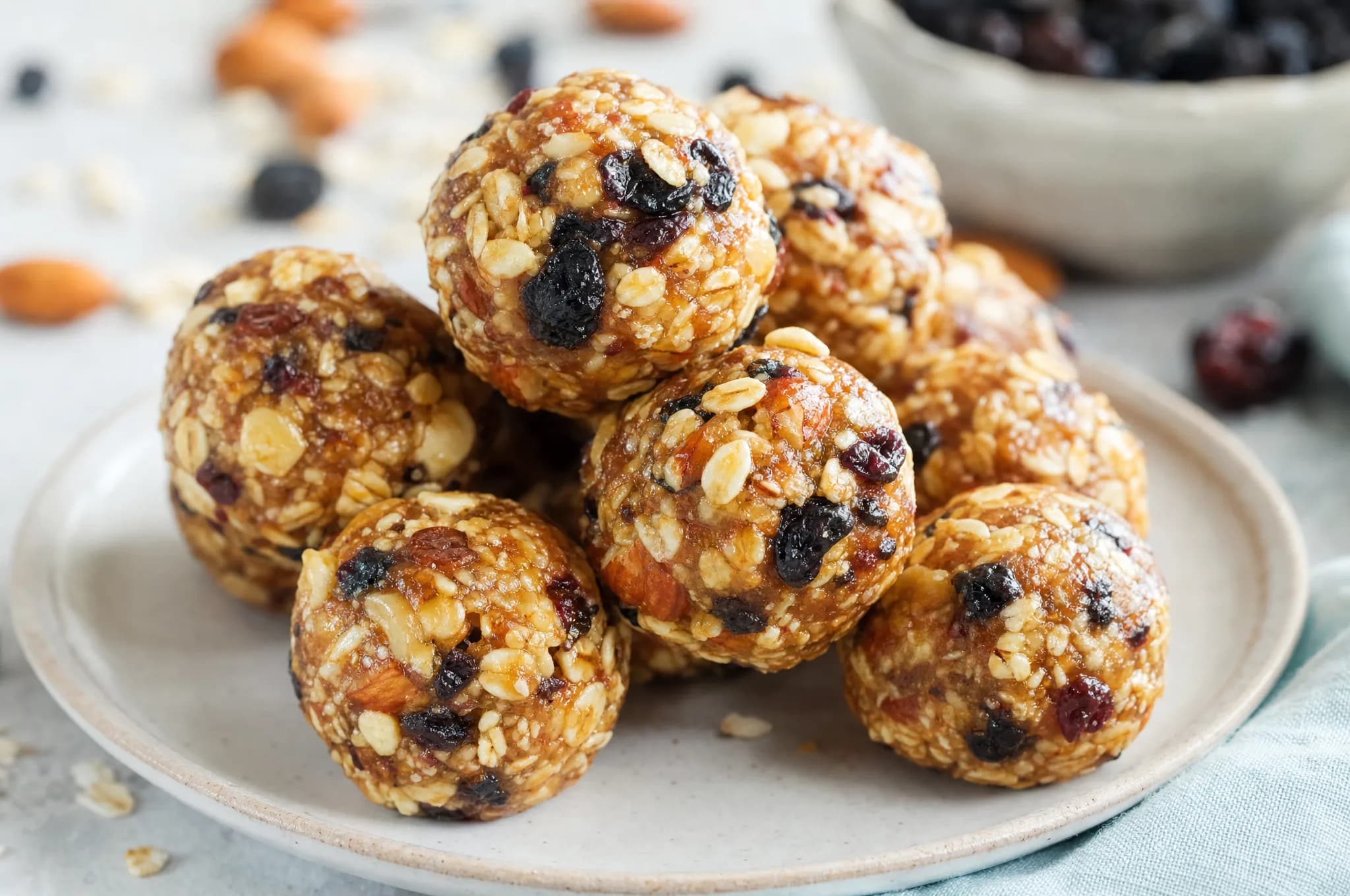 A plate of round no-bake berry-almond energy bites flecked with dried blueberries and chopped almonds, a few stacked and one bitten in half to show the oat interior