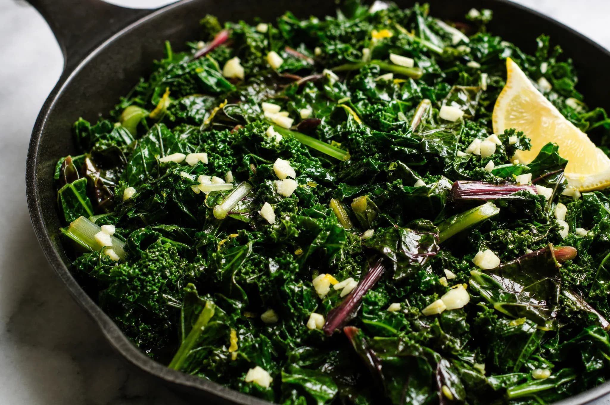 A skillet of dark leafy greens sautéed in olive oil with slivered garlic and red pepper flakes, glossy and wilted, a lemon wedge on the side