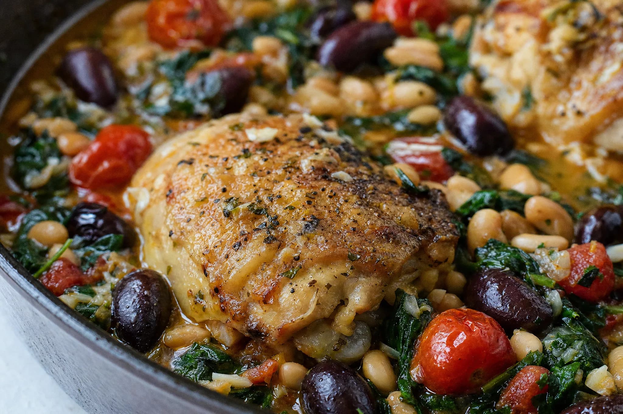 Seared chicken thighs simmered with cherry tomatoes, kalamata olives, and cannellini beans with wilted spinach in a skillet, drizzled with olive oil
