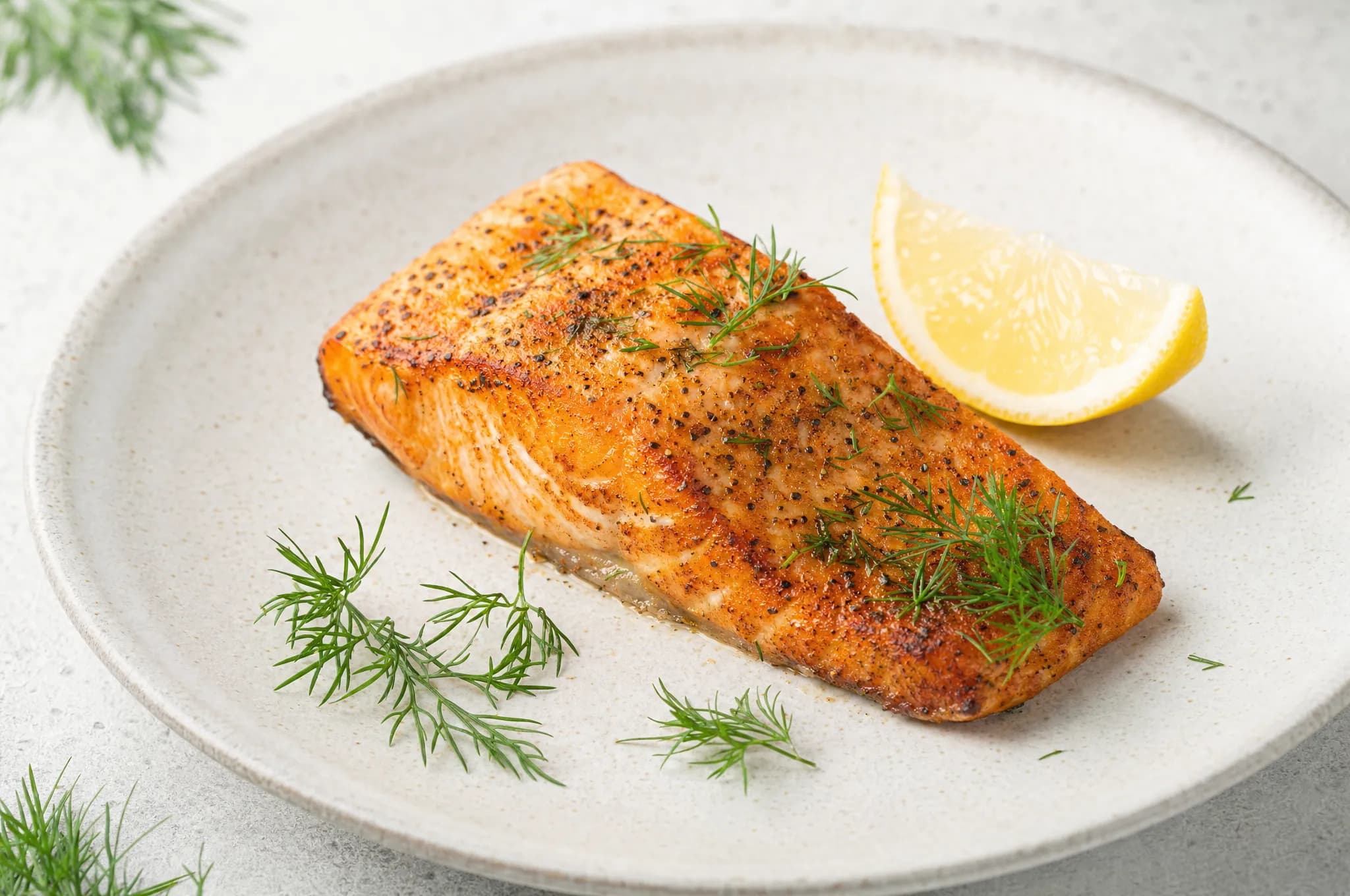 An air fryer salmon fillet with crispy edges, fresh dill, and a lemon wedge on a pale stoneware plate