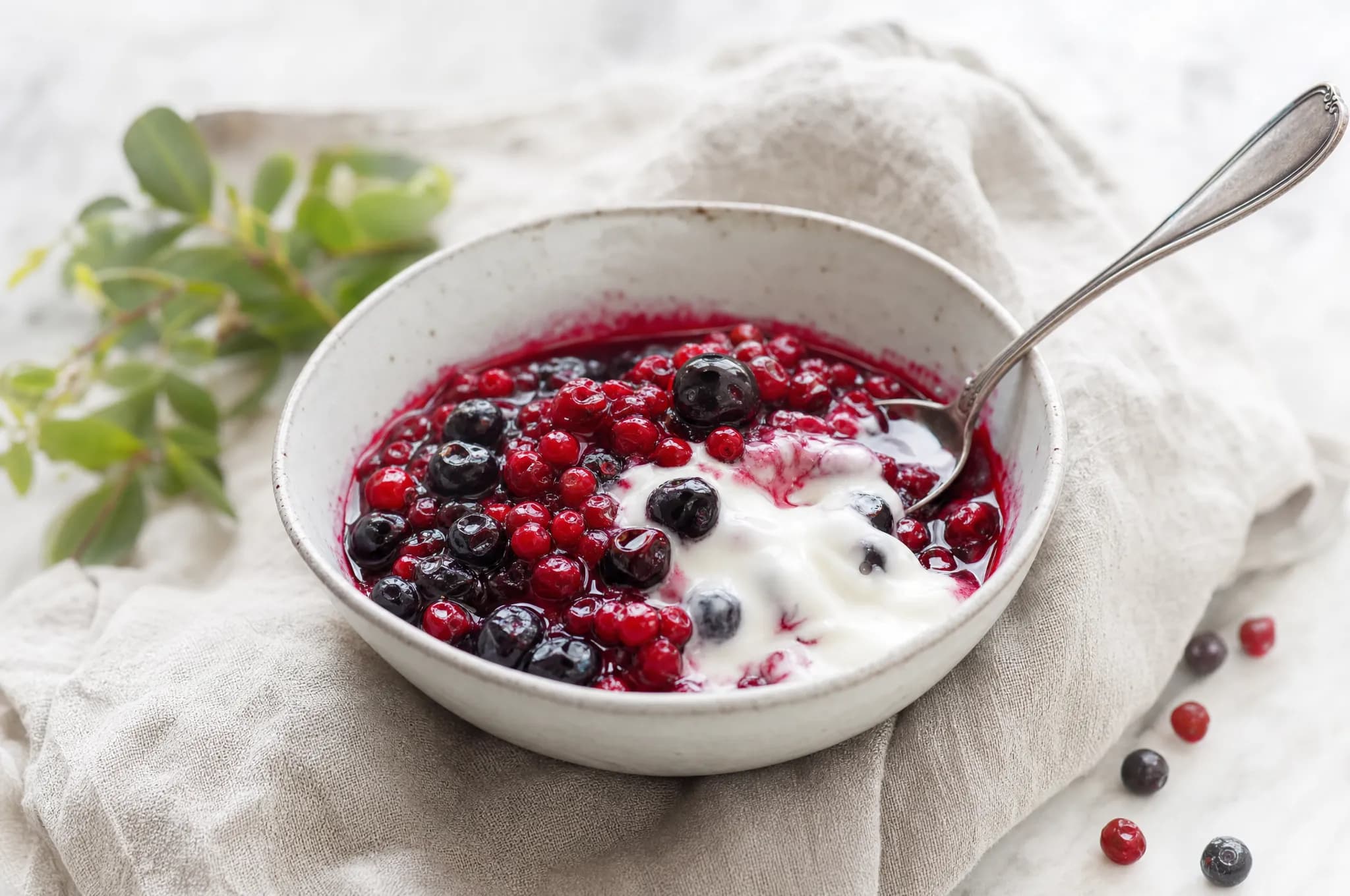 A small bowl of deep red Nordic berry compote with whole berries and a silver spoon, on a pale linen cloth