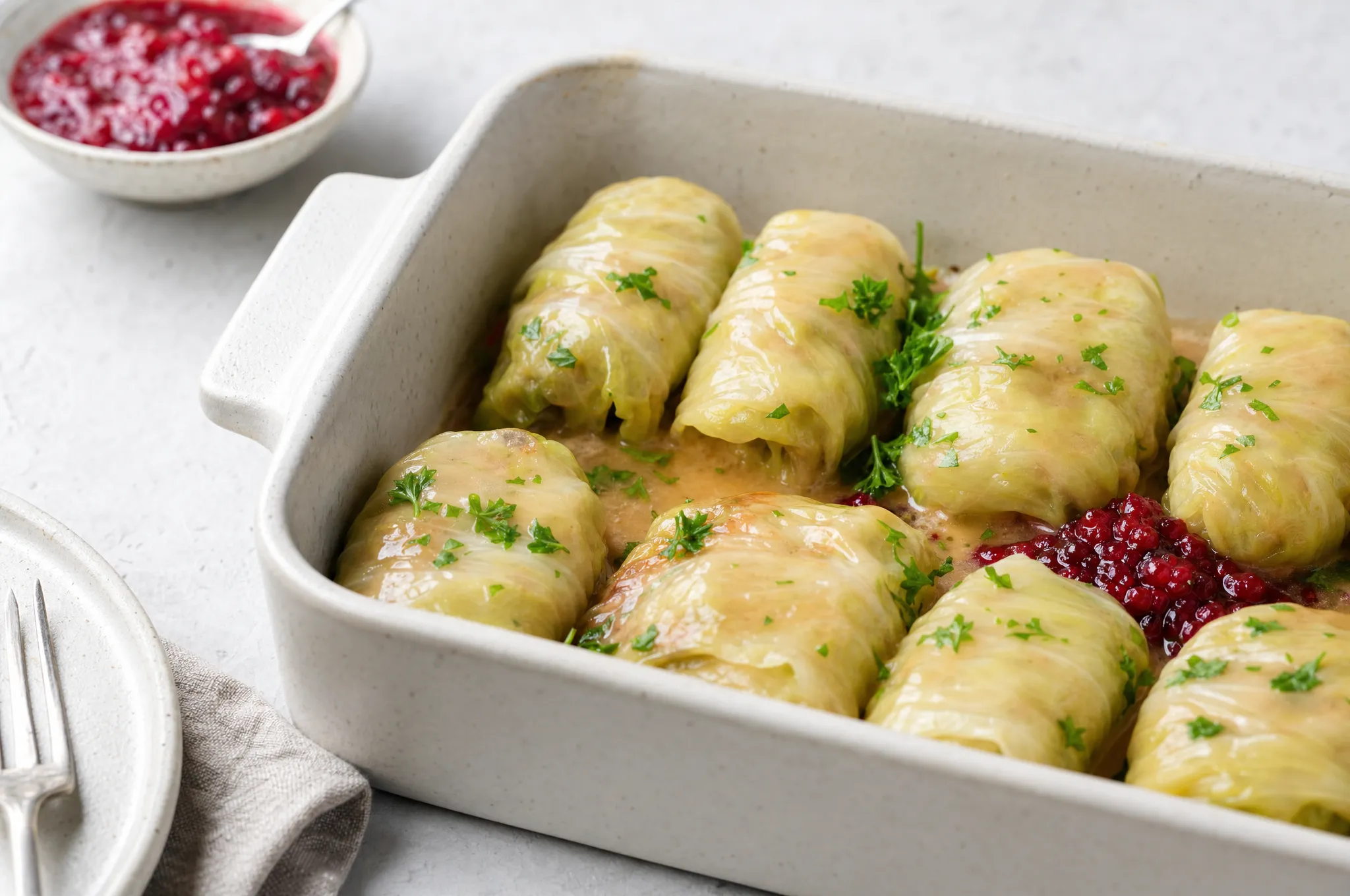 Swedish cabbage rolls in a baking dish with a light pan gravy, a spoon of lingonberry, and fresh parsley