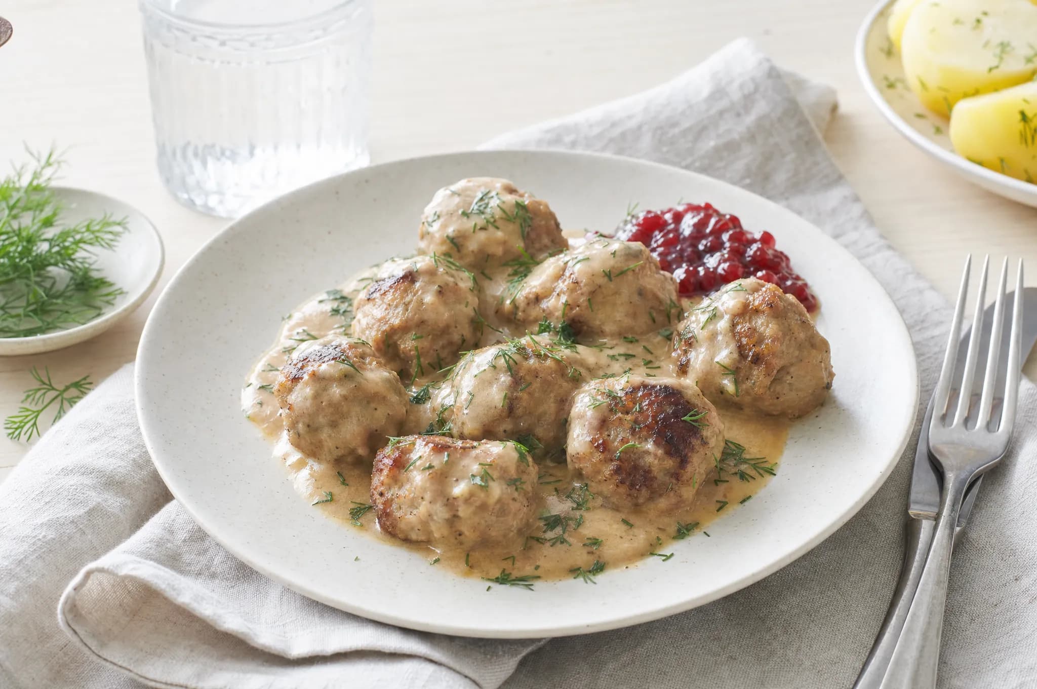 Swedish meatballs in creamy gravy with a spoon of red lingonberry jam and fresh dill on a pale stoneware plate