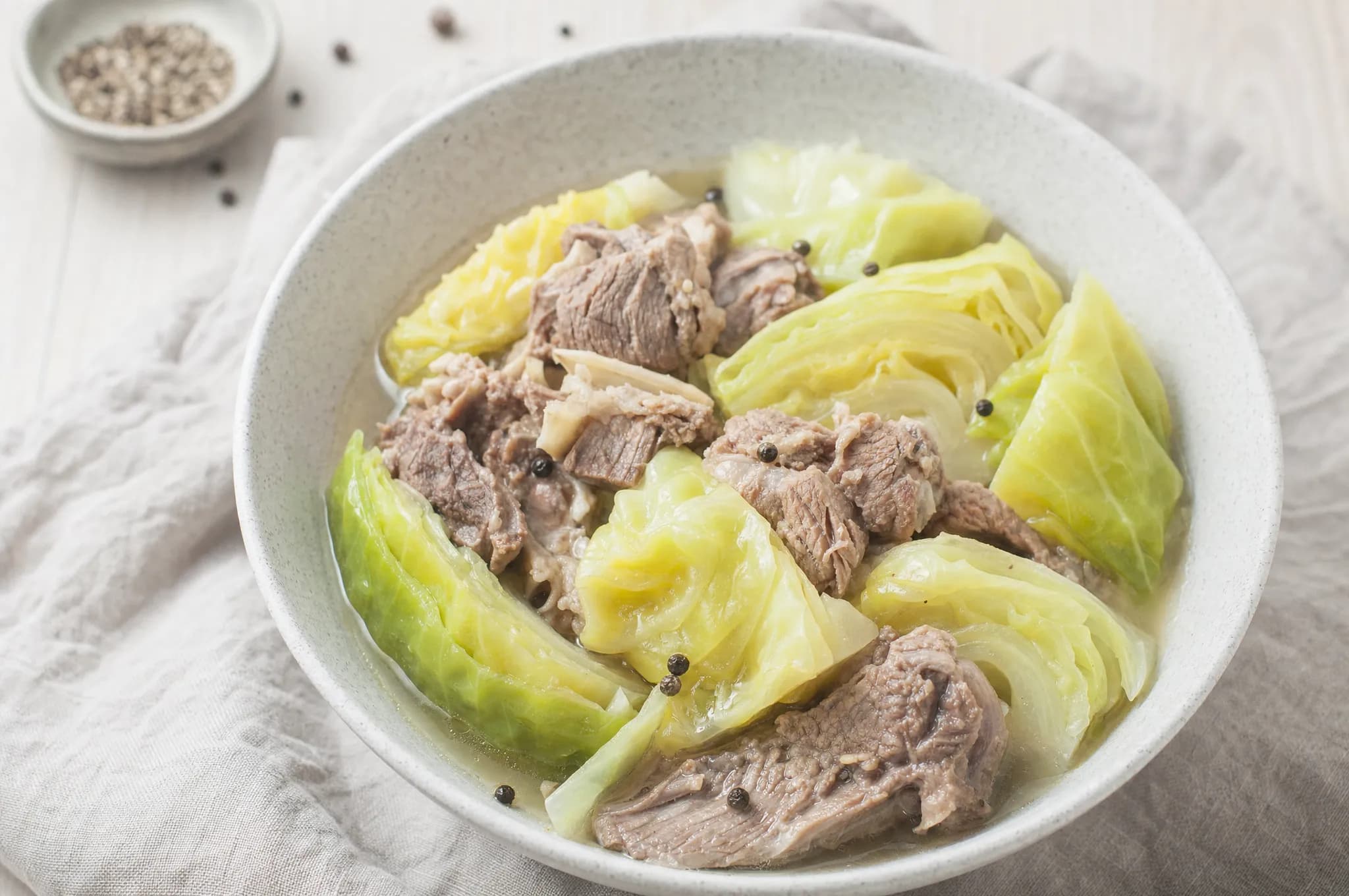 Norwegian lamb and cabbage stew (fårikål) — tender lamb with wedges of green cabbage and whole peppercorns in broth in a pale stoneware bowl