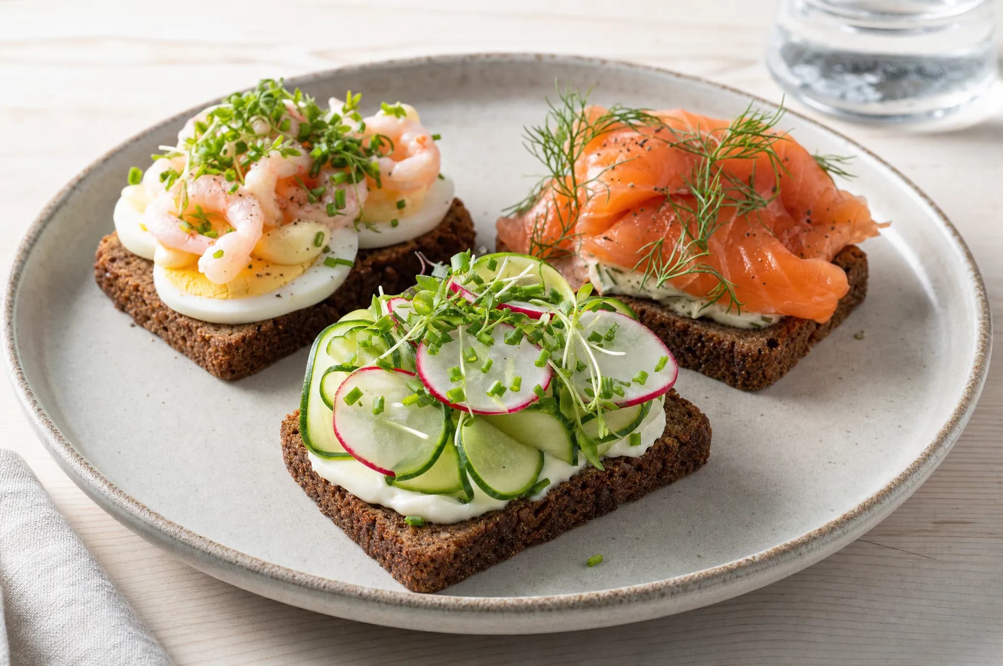 Three open-faced dark rye sandwiches topped with egg and shrimp, smoked salmon and dill, and cucumber and radish on a stoneware plate