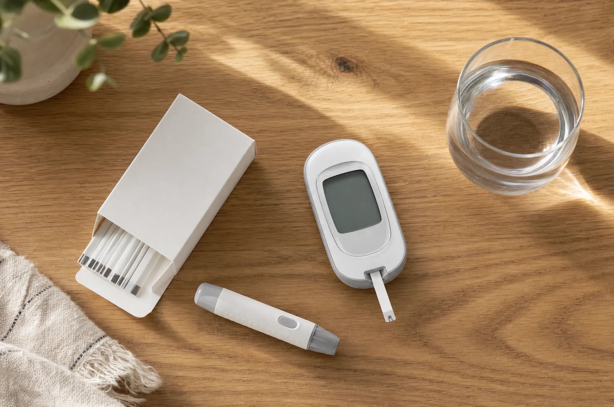 A blood ketone meter with a test strip inserted, a box of test strips, a lancet pen, and a glass of water on a sunlit wooden counter