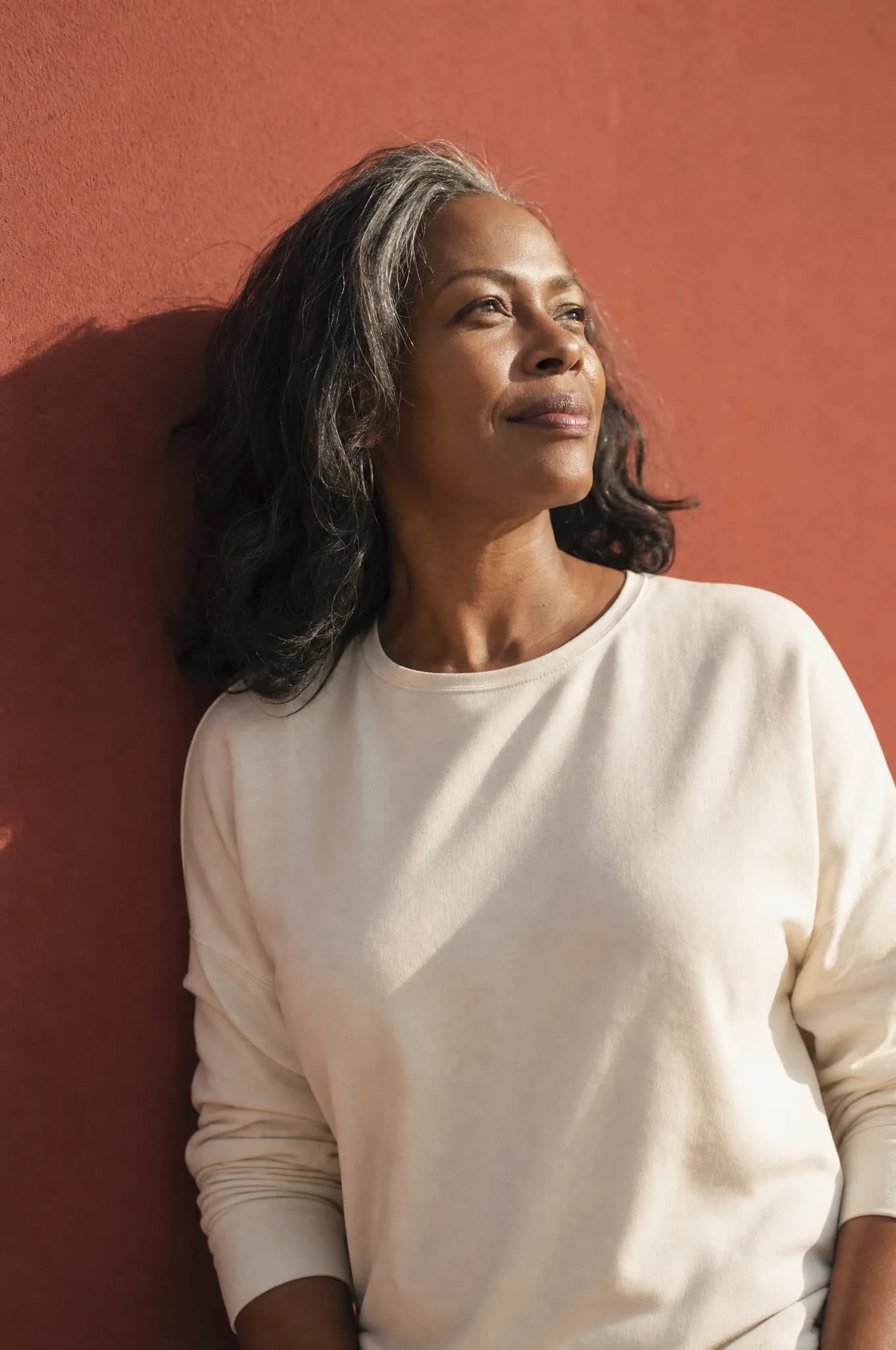 A woman with silver-streaked hair in a cream top against a coral-red wall