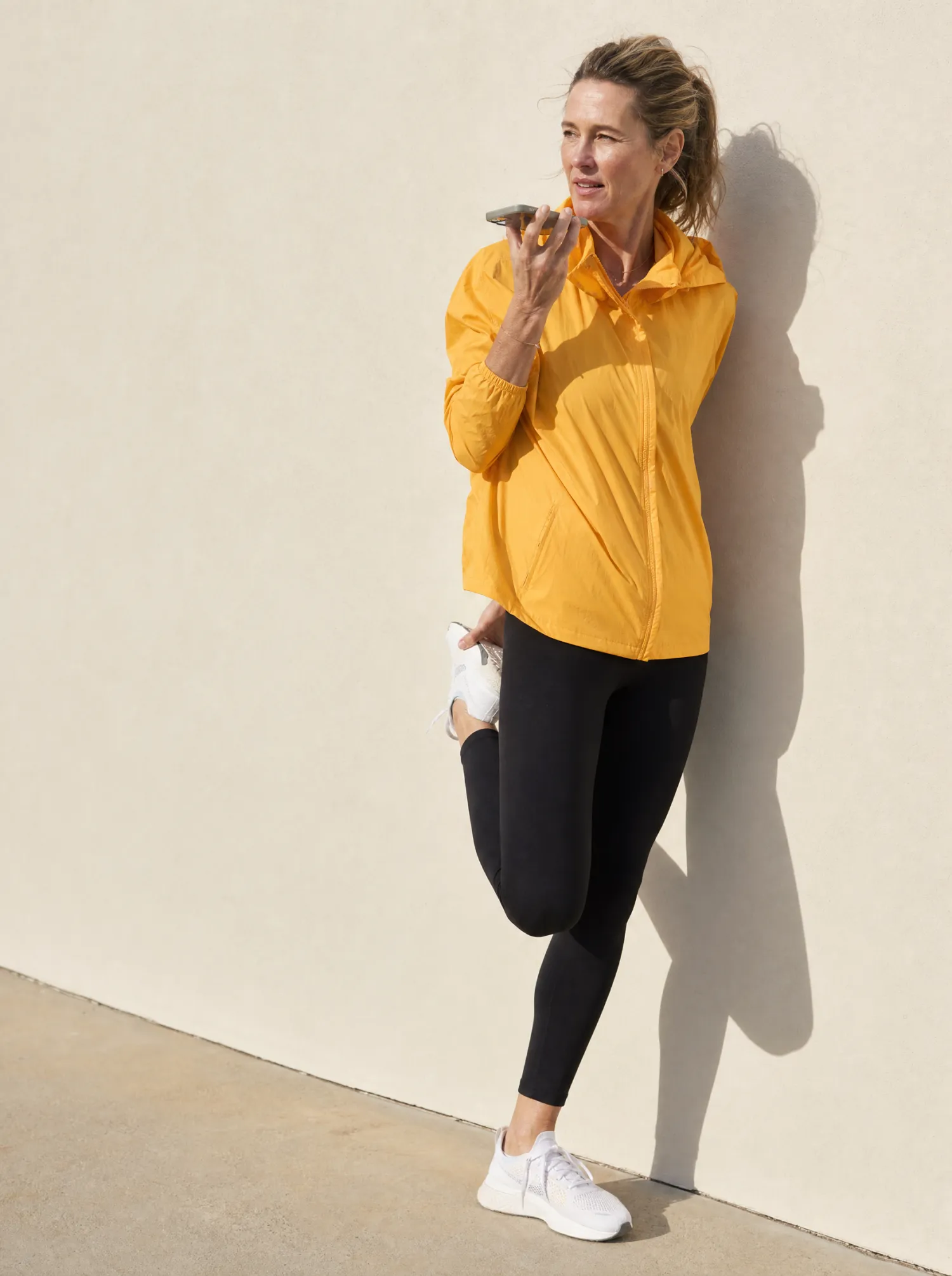 A woman in her fifties in an amber jacket and black leggings leans against a sunlit cream wall, speaking into a phone held to her mouth